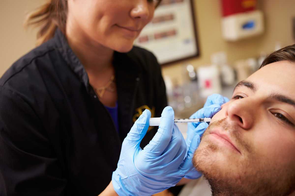 Image of a female aesthetic nurse inserting a needle into the cheek of a male patient in order to administer a cosmetic treatment