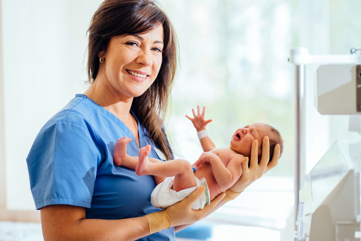 A NICU nurse holds a baby and smiles at the camera.