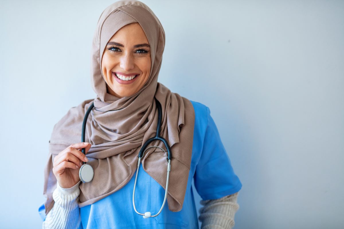 An RN wearing a hijab and holding her stethoscope smiles at the camera.