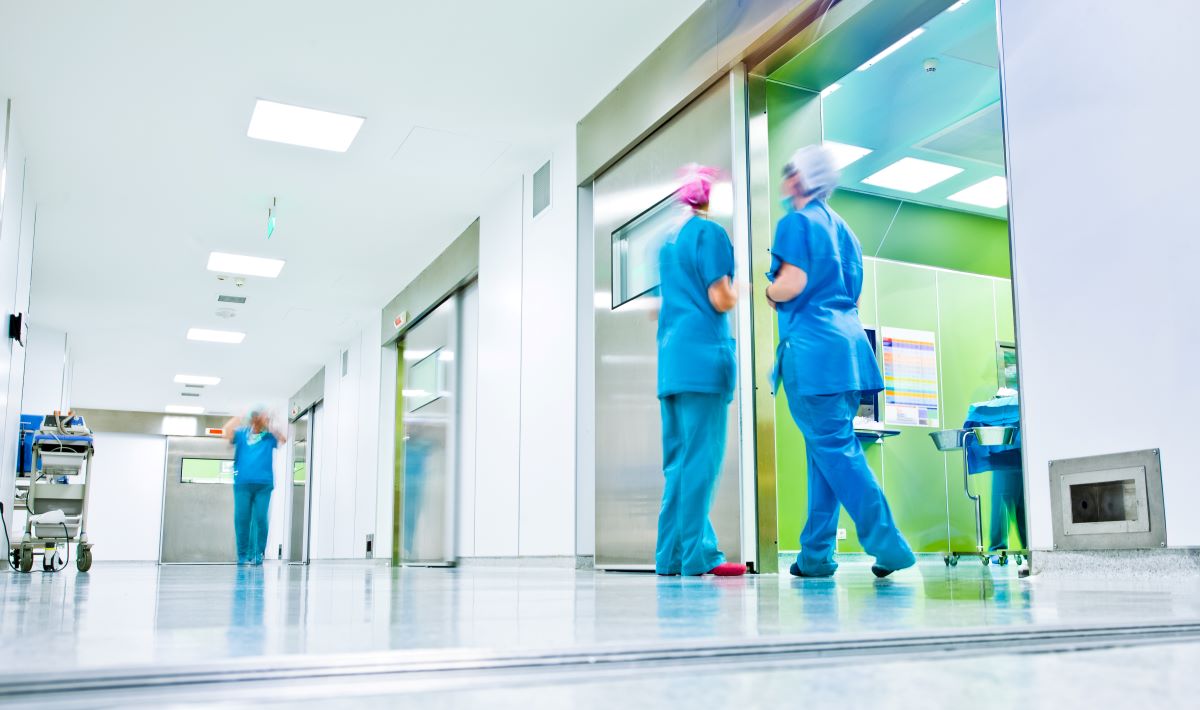 Wide shot of a hospital hallway, showing a few nurses.