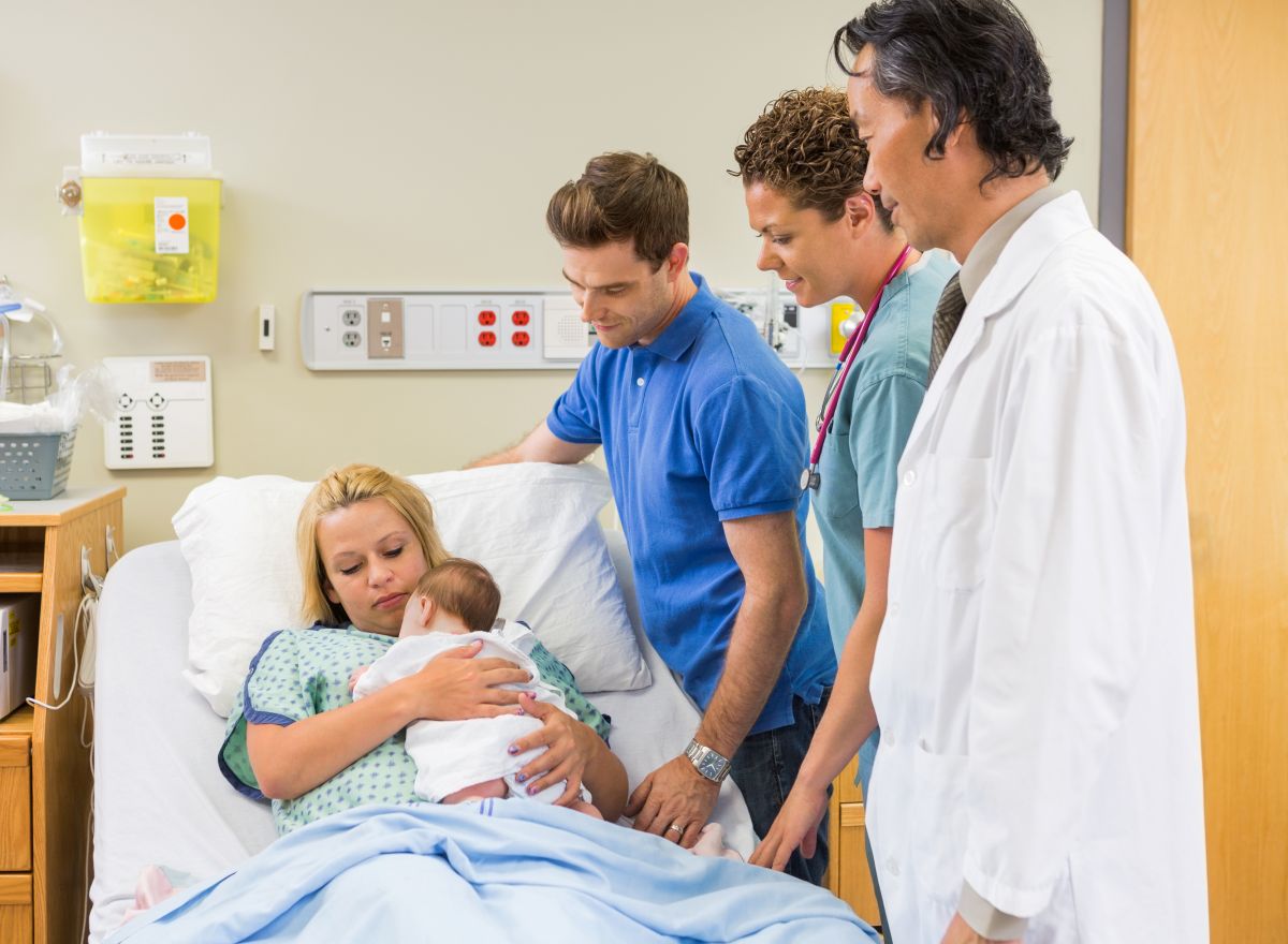 A new mom holds her baby as her partner, the doctor, and postpartum nurse look on.