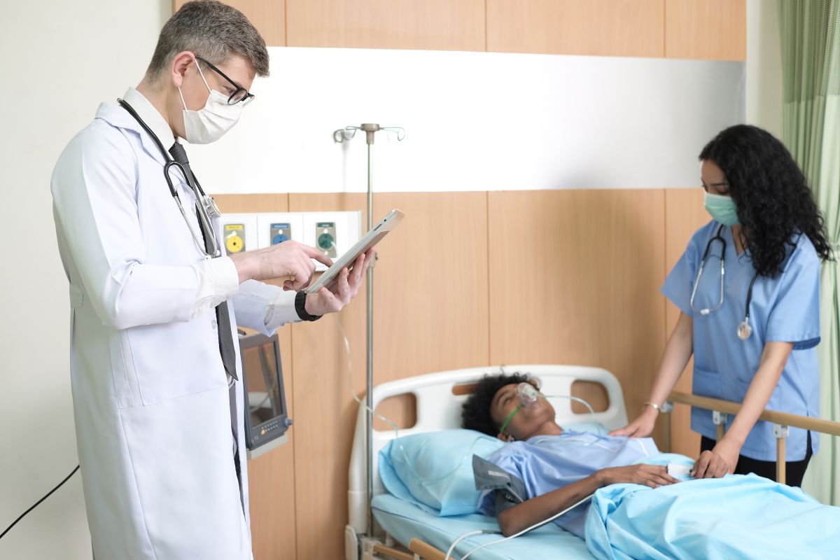 A critical care nurse assists a patient while a physician checks their chart.